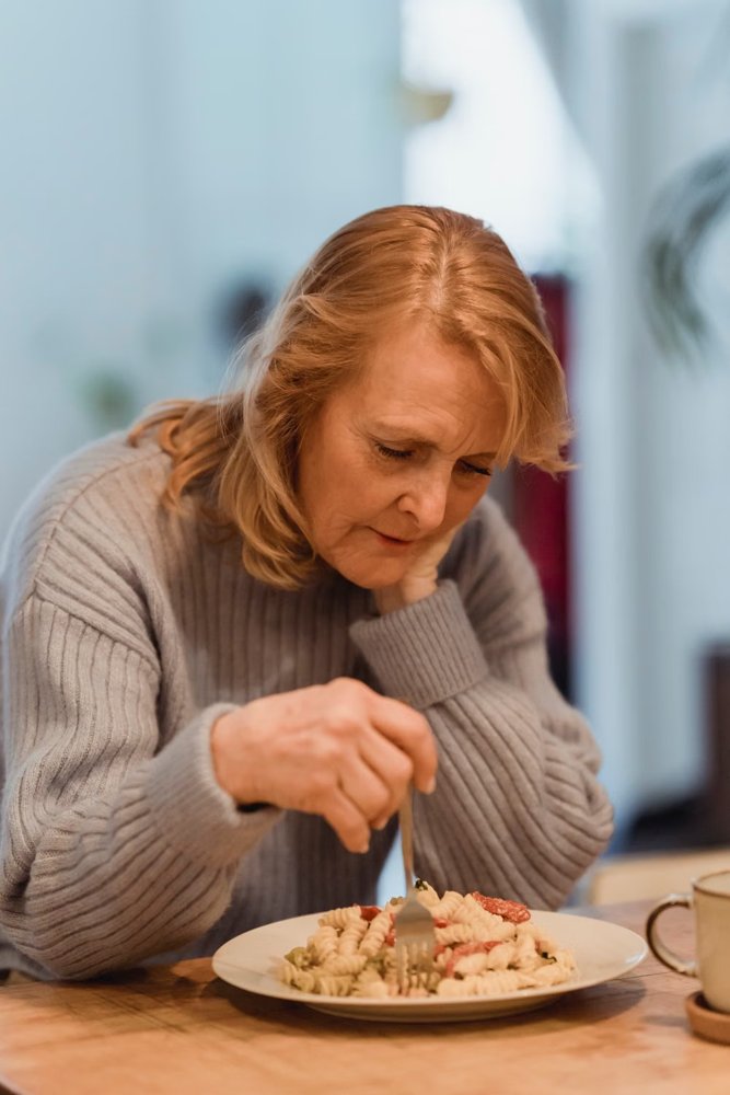 Older woman sitting at a kitchen table with a plate of pasta